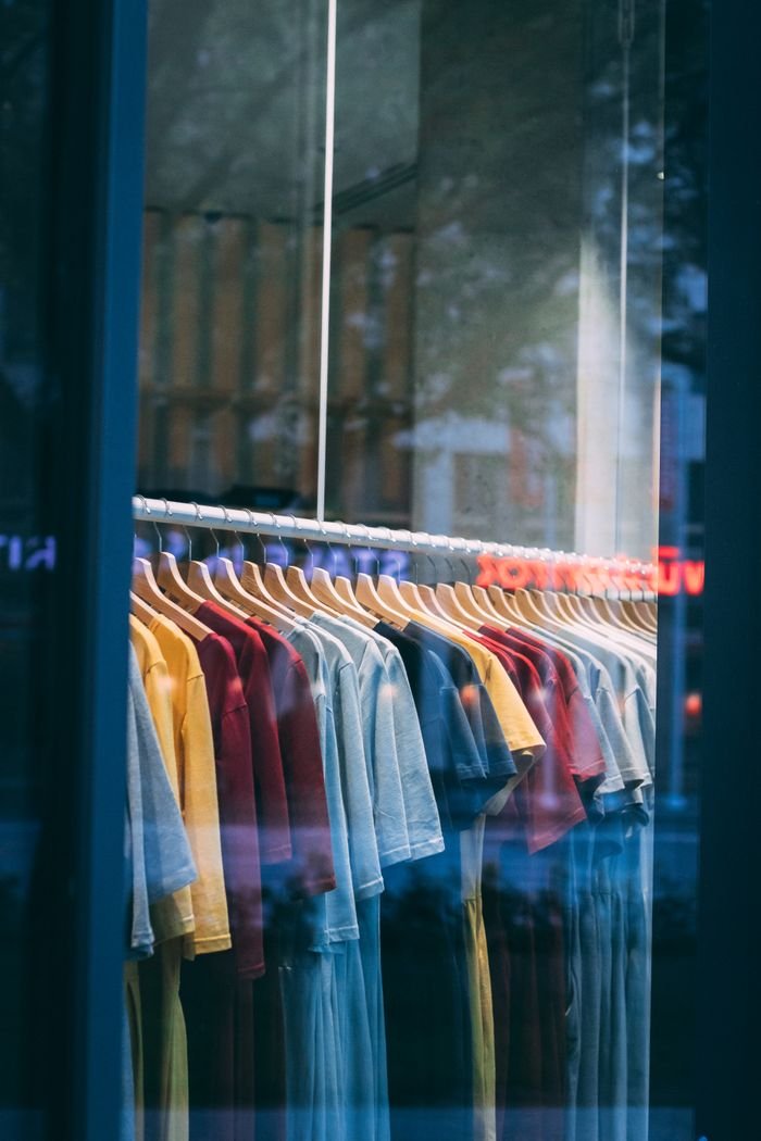 social-feed-img-03 Colorful t-shirts hanging in a store window, showcasing a vibrant display.