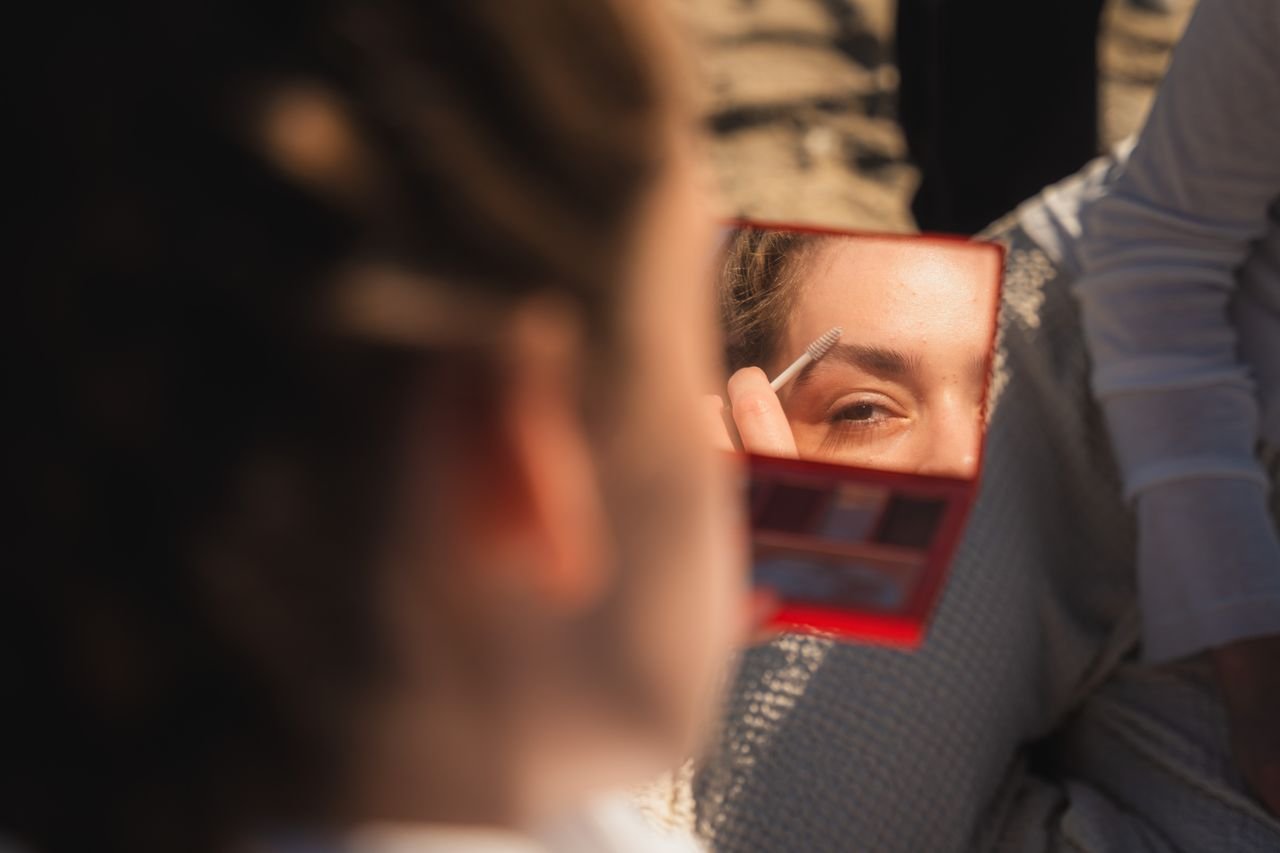 A woman uses a mirror to apply eyebrow makeup outdoors, captured in warm natural light.